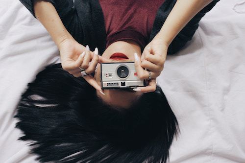 Woman lying on bed holding camera for selfie, dark hair, casual outfit.