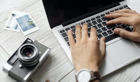 Person typing on a laptop with camera and photos on a wooden desk.
