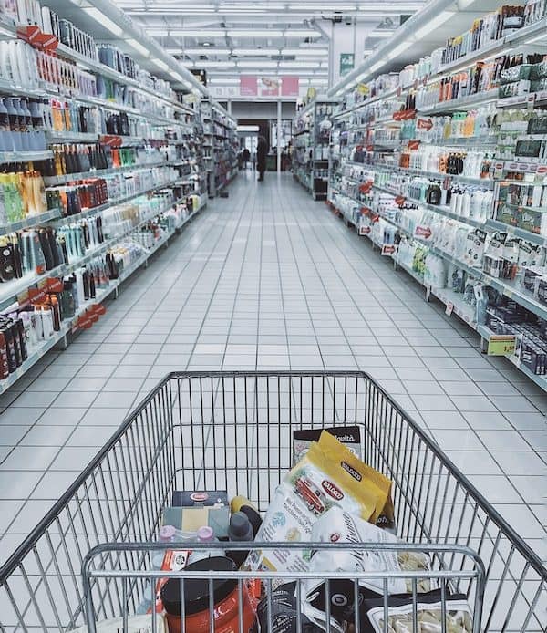Wide view of a grocery store aisle with shopping cart filled with products.