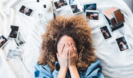 Woman with curly hair covering face surrounded by instant photos and camera on bed.