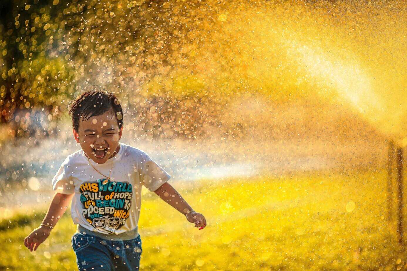 A young boy running through a water sprinkler wit the sun in te background
