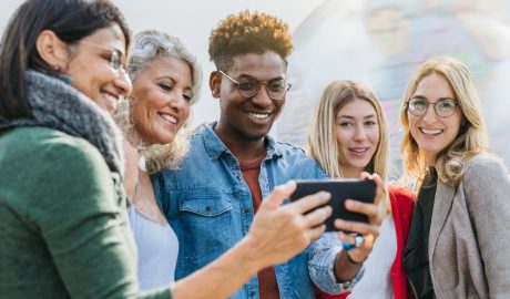 A group of smiling people of varying ages and ethnicities sharing a moment around a smartphone.
